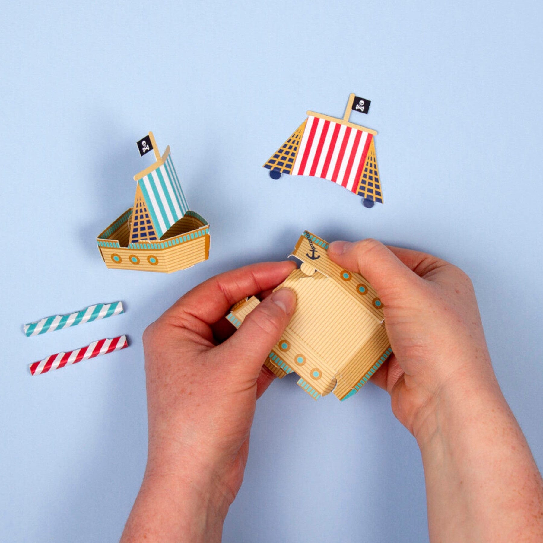 Hands assembling paper craft pirate ships with striped sails on a blue background