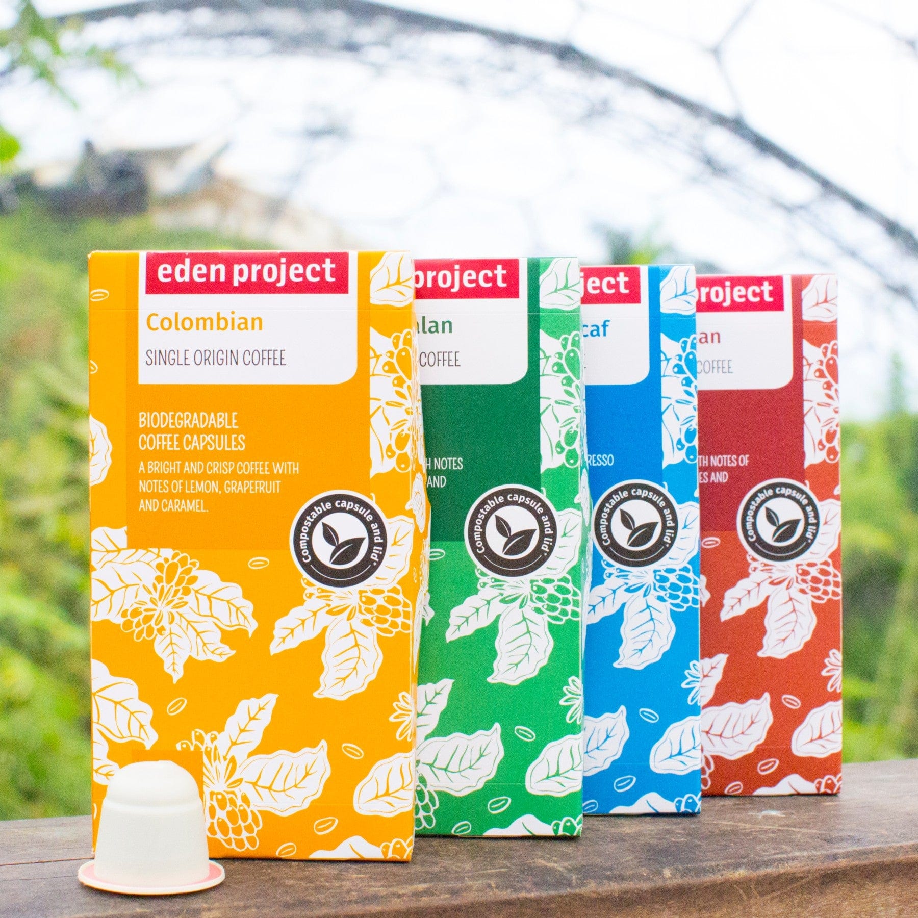 Four colorful packages of Eden Project biodegradable coffee capsules with labels indicating Colombian, Guatemalan, Decaf, and Peruvian varieties, displayed outdoors on a wooden surface with green foliage in the background.