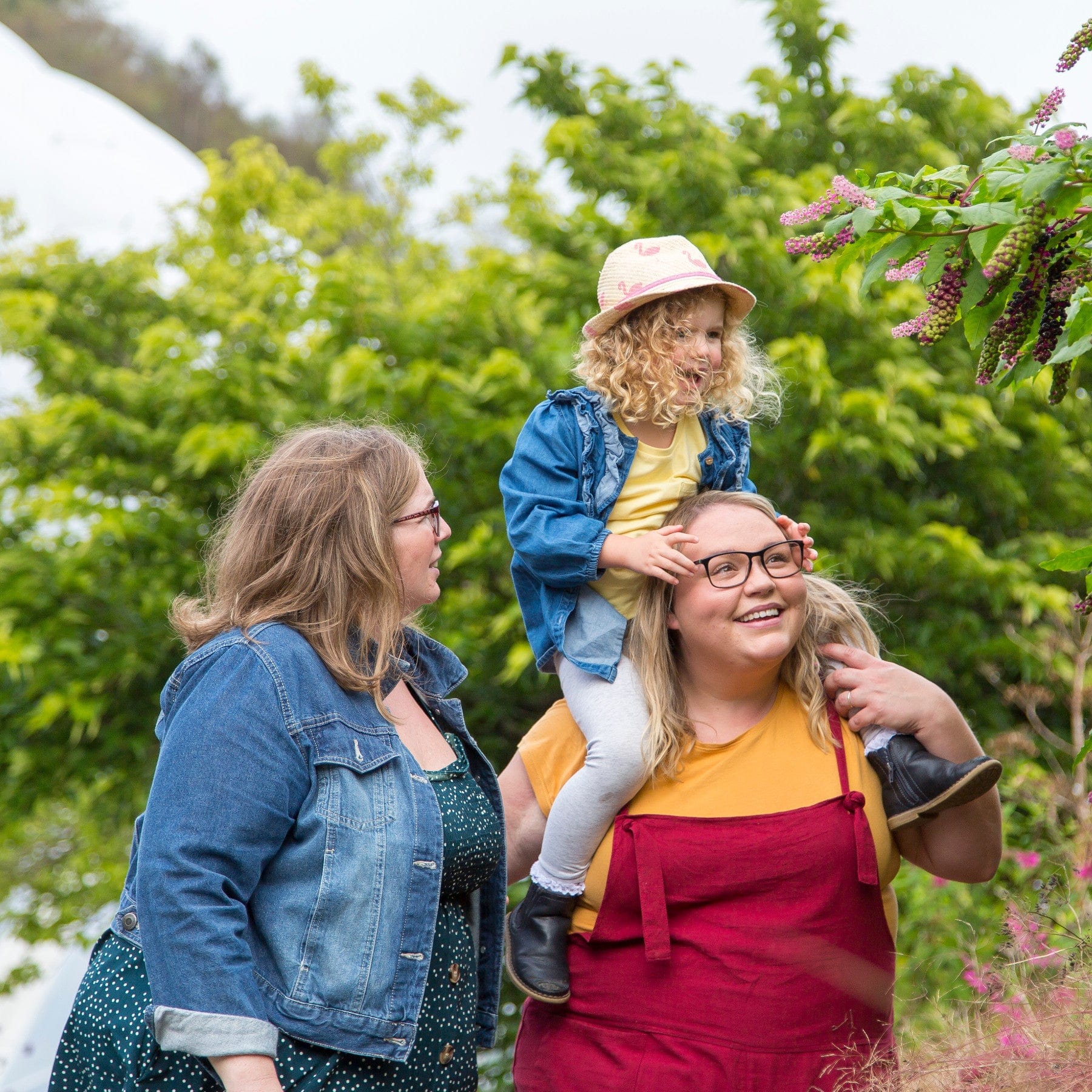 Family enjoying outdoor time together with a woman smiling at a child on her shoulders, another woman looking at them, greenery in the background, casual clothing, happy family moment, nature setting, bonding, and togetherness.