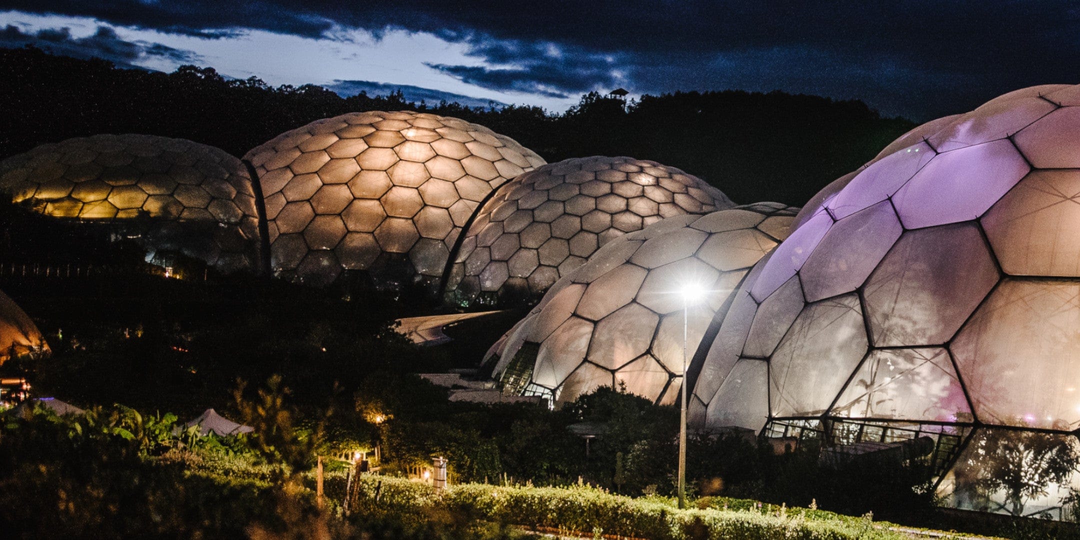 Night view of illuminated geodesic domes at eco-park, hexagonal patterns on futuristic structures, surrounded by darkened natural landscape under evening sky.