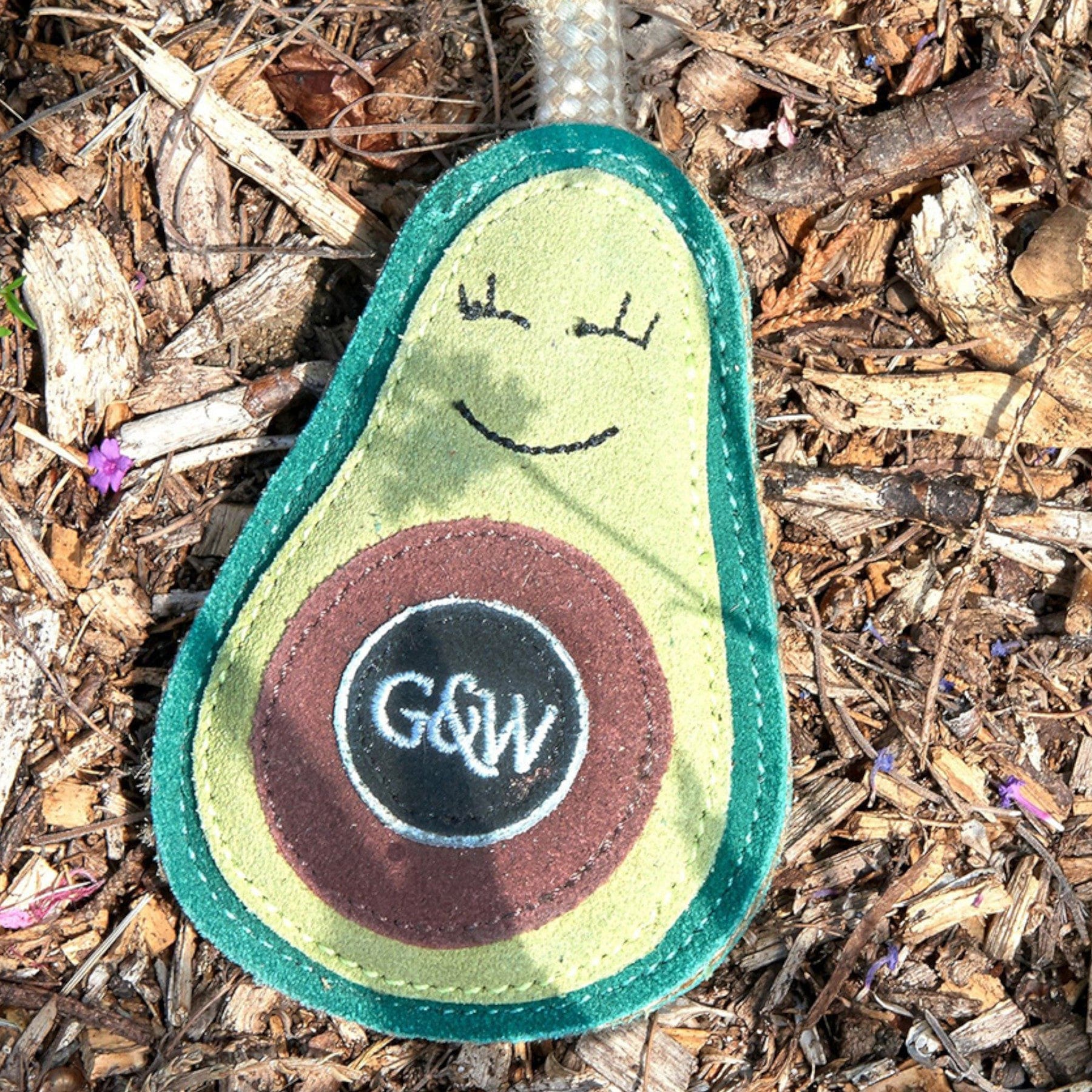 Alt text: Smiling avocado plush toy with a happy face on a bed of wood chips and twigs, featuring vibrant green and brown colors with a prominent seed design displaying "GW" initials.