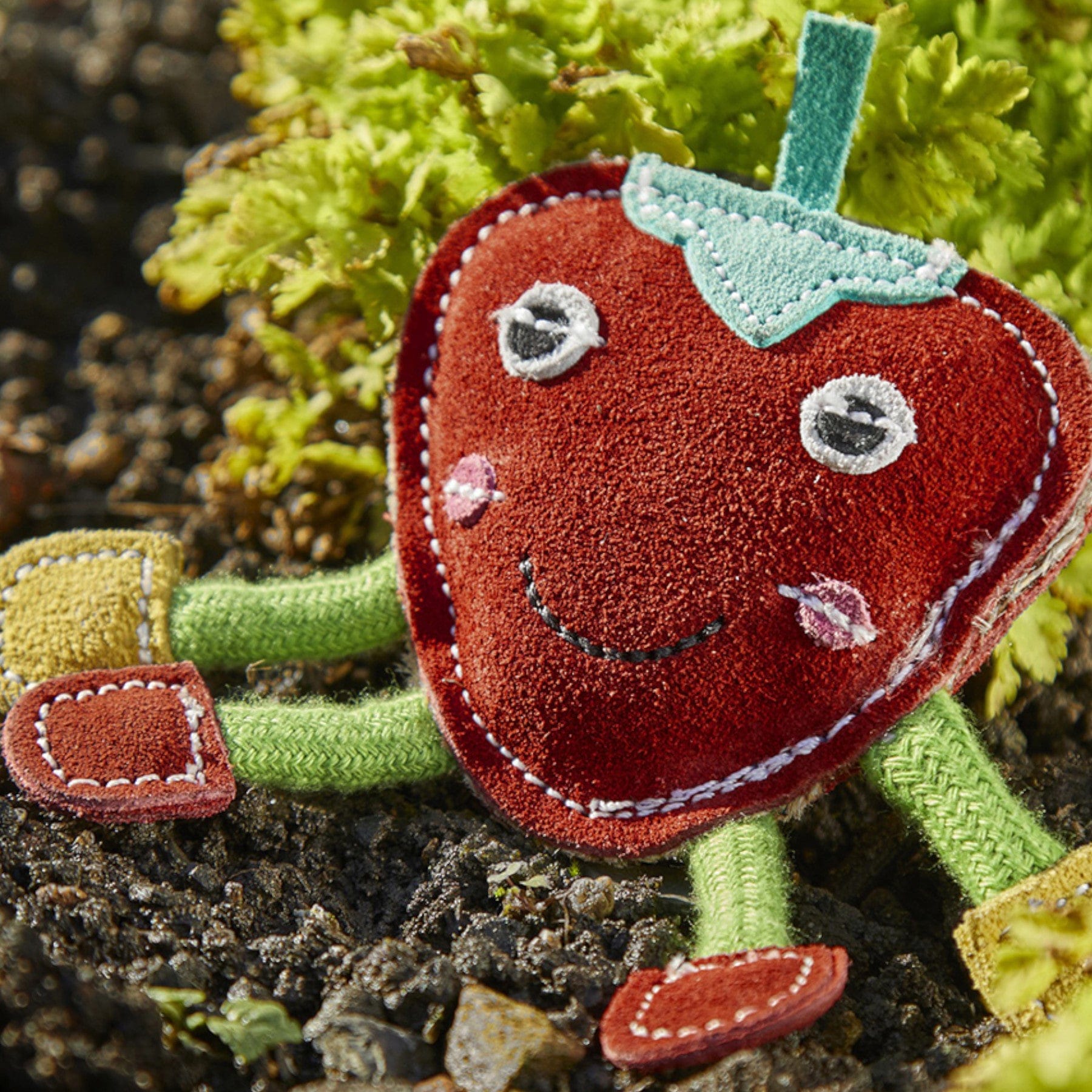 Alt text: Plush toy of a smiling strawberry character with arms and legs, sitting in a garden with lettuce in the background, detailed embroidery, crafted for play and education.