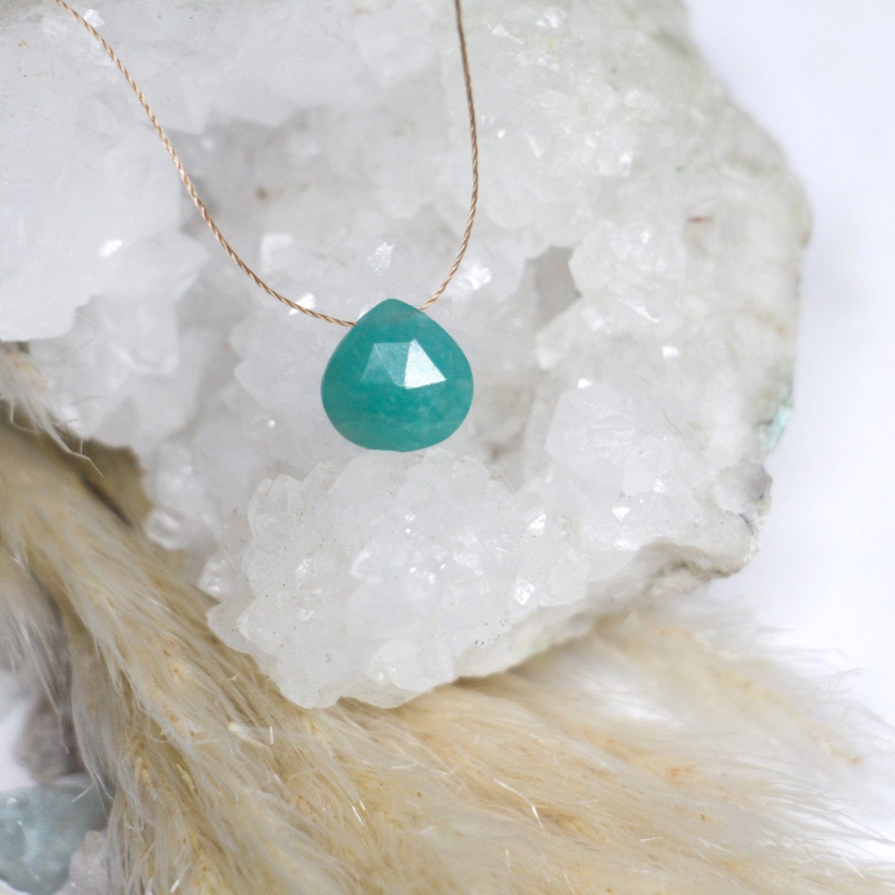 A teal faceted gemstone pendant on a delicate gold chain, displayed against a backdrop of white crystalline structures and soft fur textures.