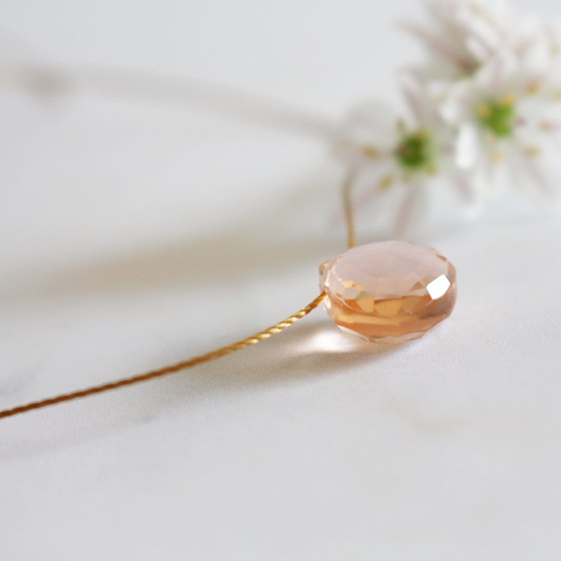 Elegant pink crystal pendant on delicate gold chain with white blossoms in soft focus background on a white surface.