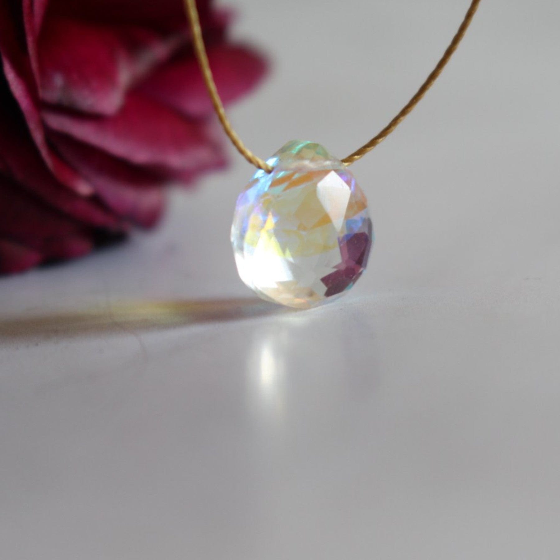 Iridescent crystal pendant on a gold necklace with a blurred rose in the background on white surface