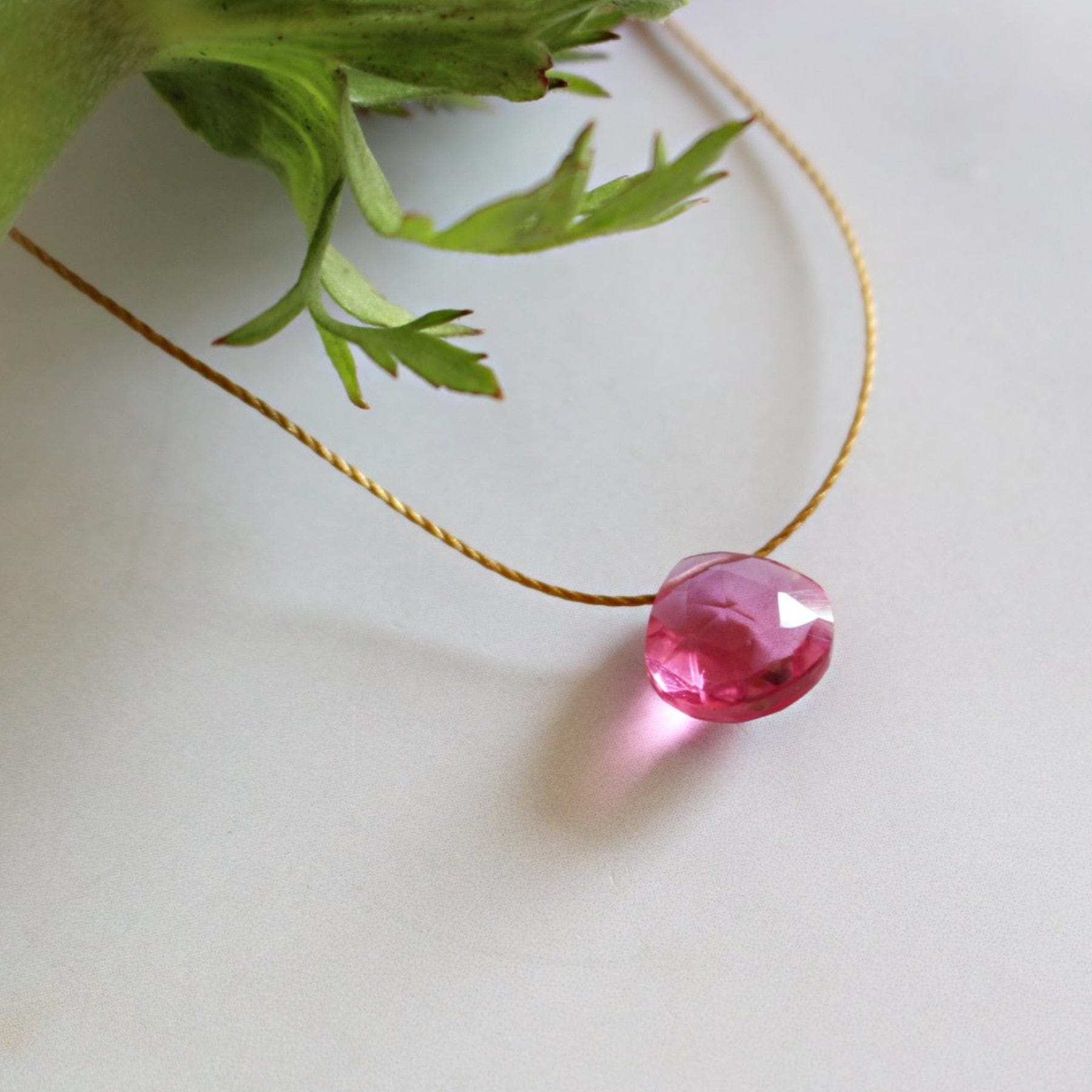 Pink gemstone pendant on gold chain next to green leafy plant on white background