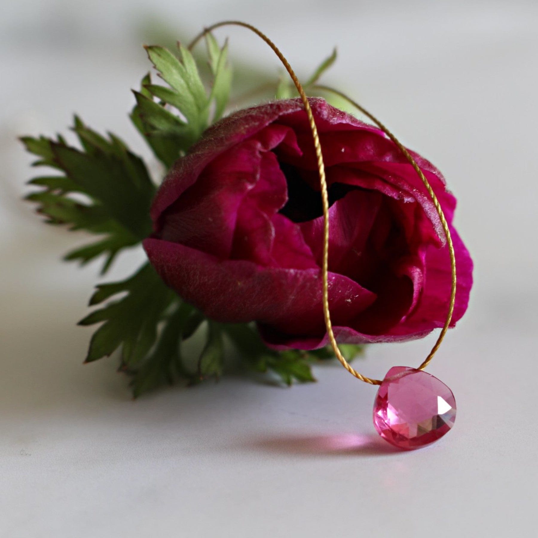 Close-up of a vibrant pink rose with a delicate golden thread through it, holding a sparkling pink gemstone against a soft-focus white background