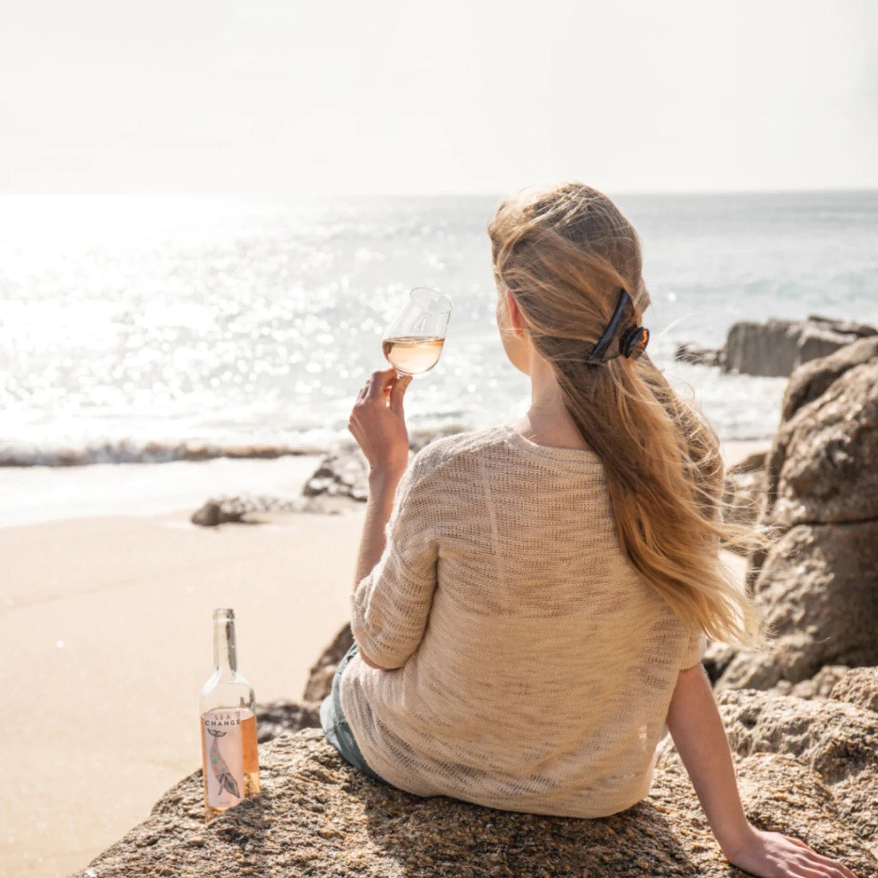 Woman sitting on beach rocks holding wine glass, ocean view, sunny day, relaxation concept, bottle of rose wine next to her