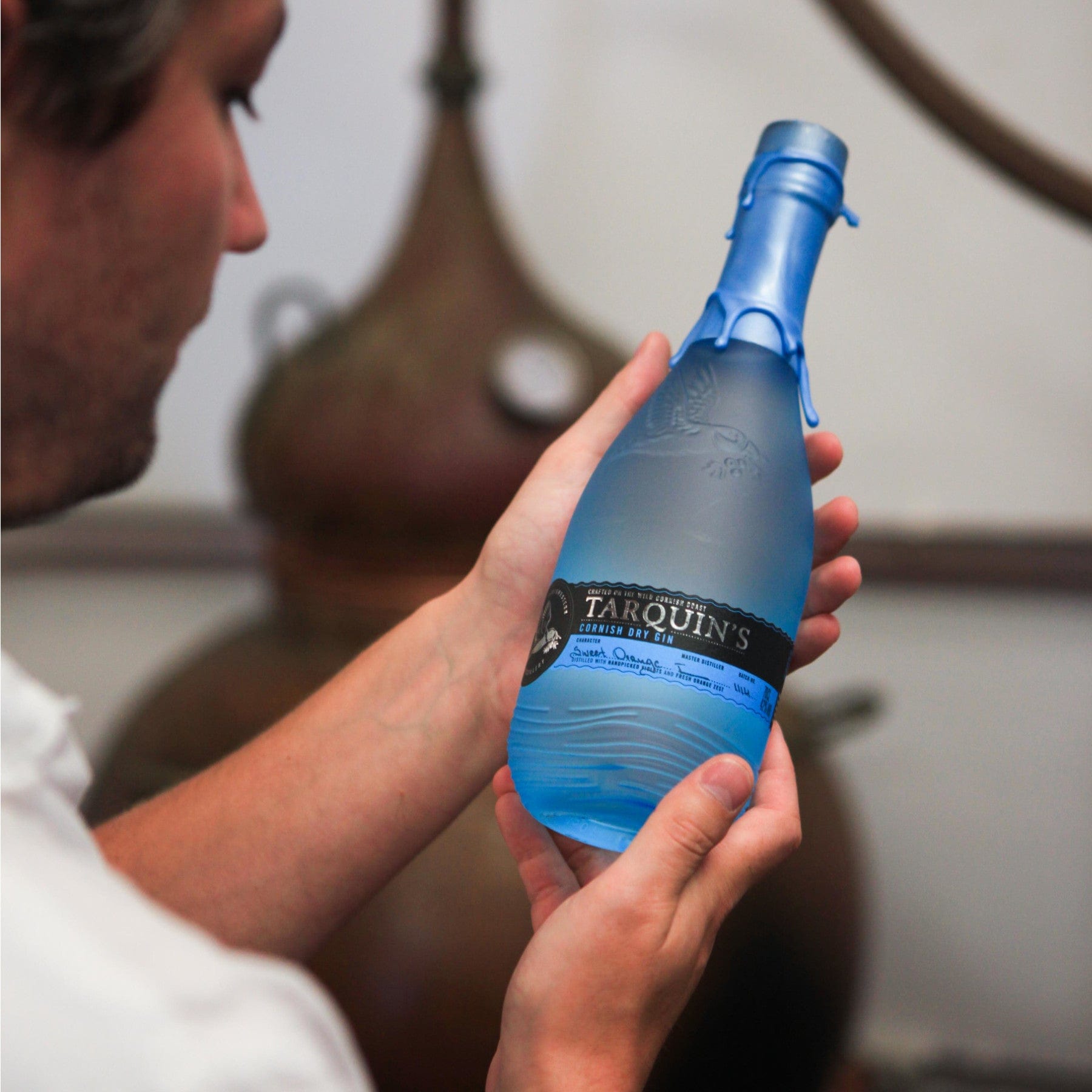 Man inspecting Tarquin's Cornish Gin bottle in distillery, blue wax seal and label, artisanal spirits, handcrafted alcohol, copper still background