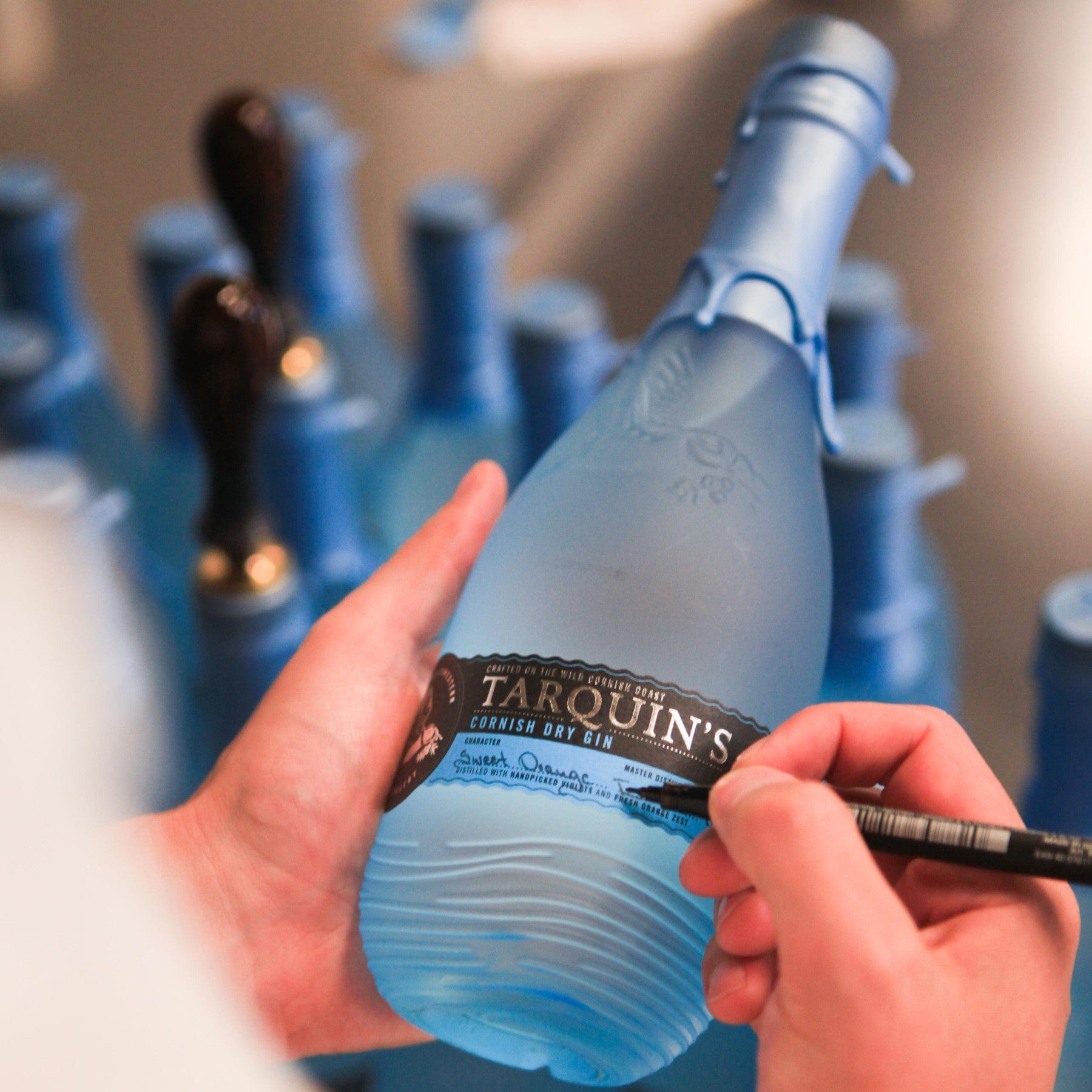 Person applying label by hand to Tarquin's Cornish Dry Gin blue bottle in a production line setting with multiple bottles in the background.