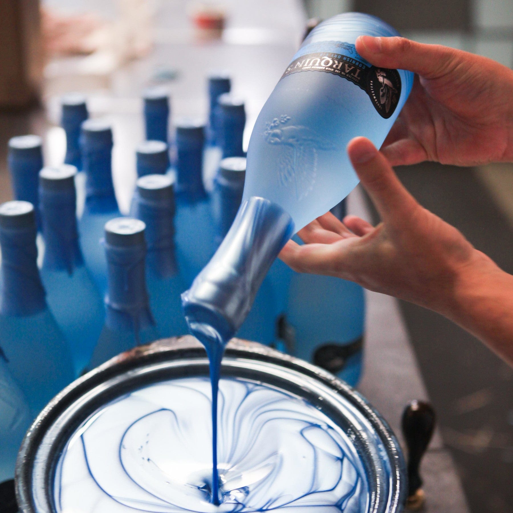 Hand pouring blue paint from a bottle into a bucket with swirling paint pattern, with blurred blue bottles in the background.