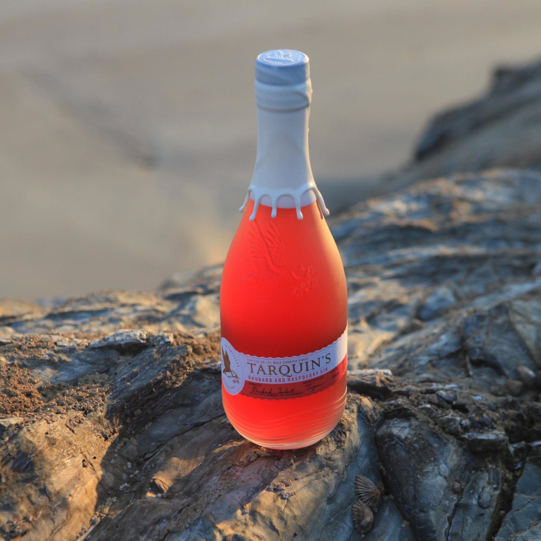 Tarquin's Cornish Gin bottle with pink hue sitting on rugged coastal rocks during sunset.