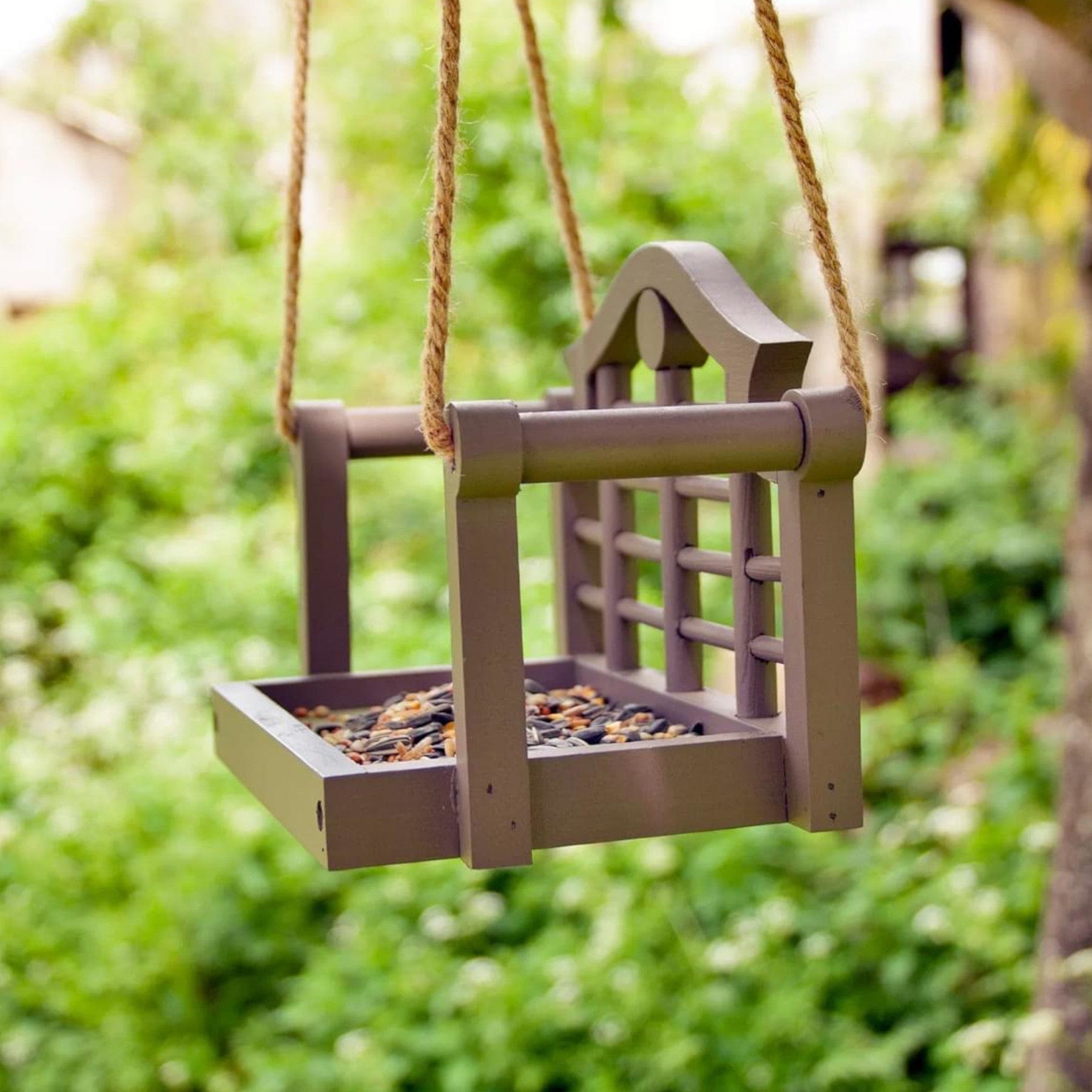 Wooden bird feeder filled with seeds hanging on ropes from a tree in a green garden setting.