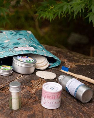 Eco-friendly toiletries on wood, including reusable pads, metal containers, toothbrush, face balm, and tooth powder with nature background