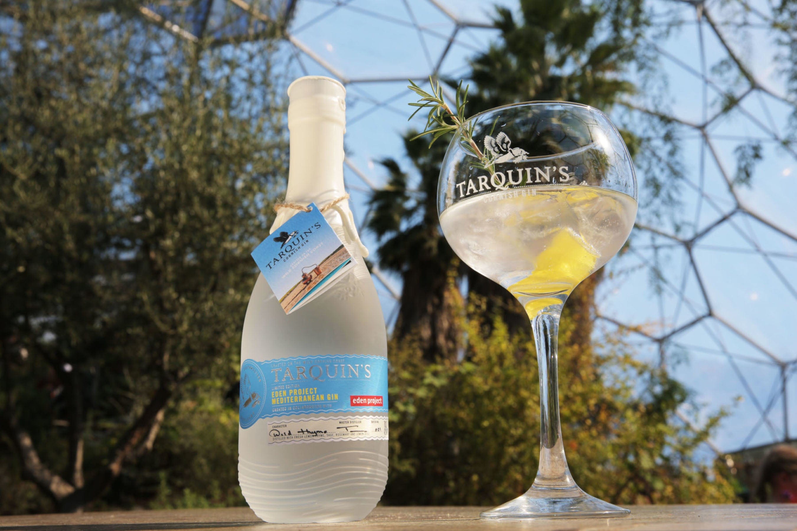 Tarquin's gin bottle with label and gin tonic cocktail in balloon glass garnished with lemon and rosemary, outdoor setting with foliage and geodesic dome in background.