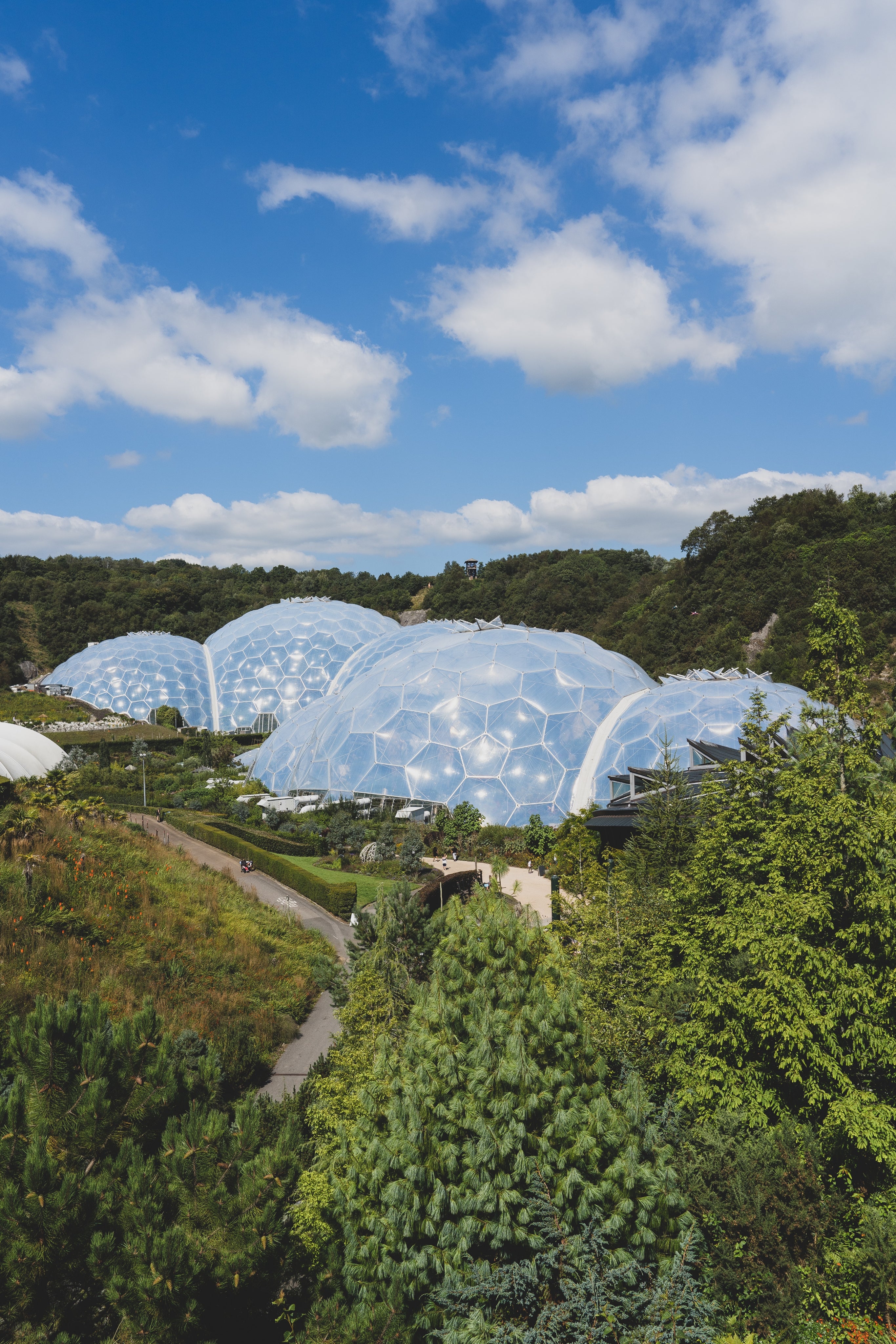 Biome conservatories with geodesic domes amid lush green landscape under a blue sky with scattered clouds.