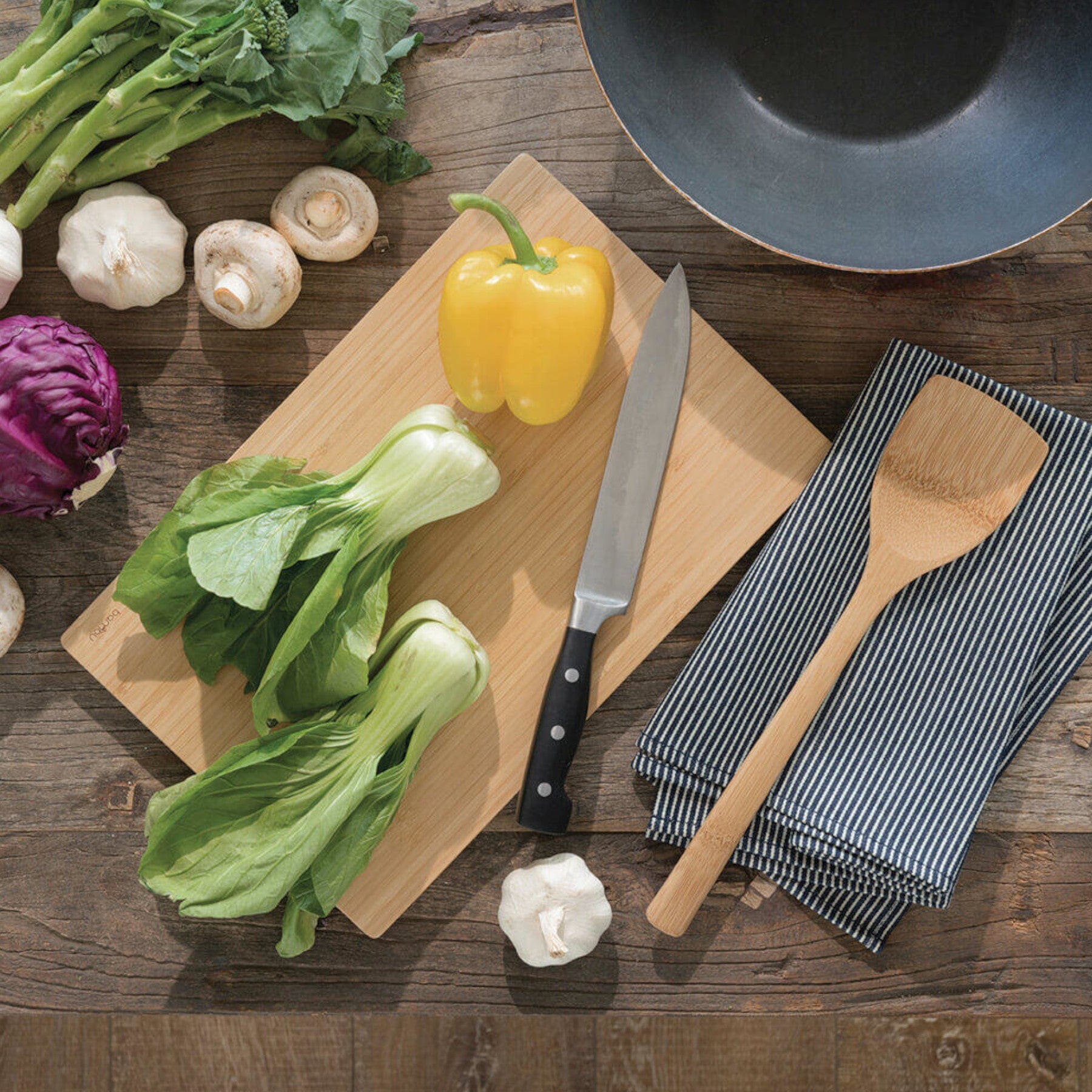 Fresh vegetables on wooden cutting board with chef's knife, wooden spoon, striped kitchen towel, and empty black pan on rustic wood tabletop, ingredients for healthy cooking and meal prep concept.