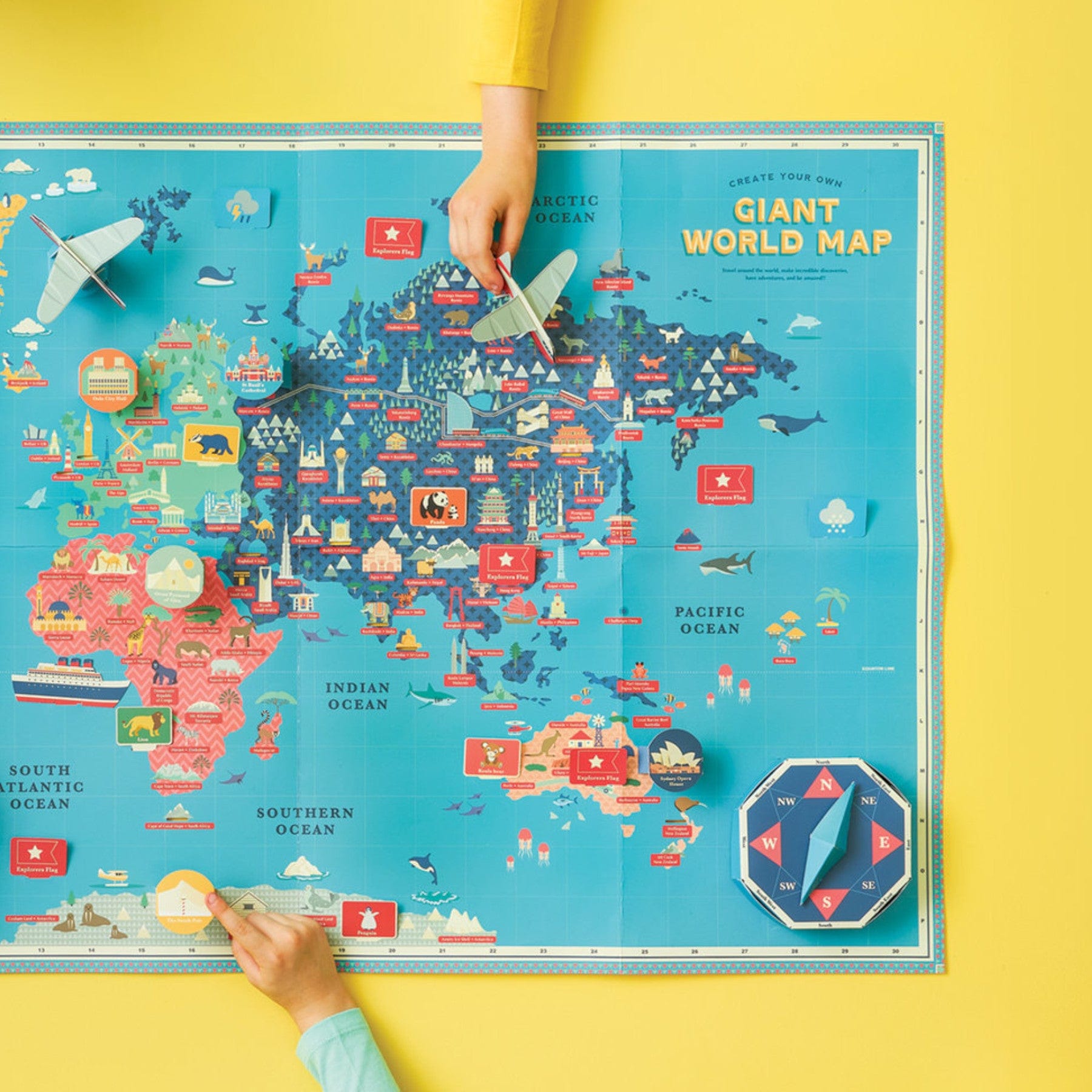 Person interacting with a colorful illustrated giant world map on yellow background, educational tool for children, geography learning activity, interactive wall map with landmarks and animals, hands placing or pointing at elements on the map.