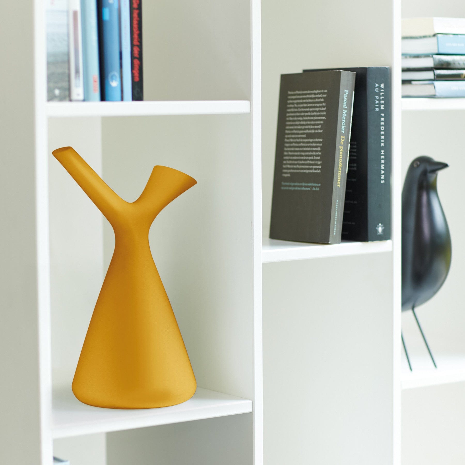 Modern yellow vase on white bookshelf with assorted books and decorative black bird figurine in a bright interior.