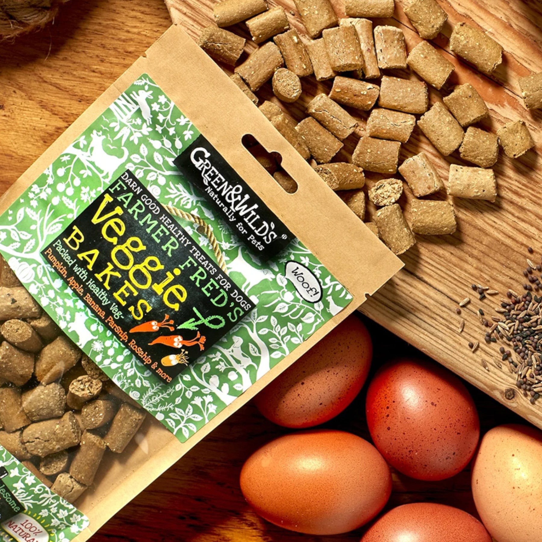 Green & Wilds Farmer Fred's Veggie Bakes dog treats packaging with loose treats scattered on wooden surface, eggs and sprinkle of seeds alongside.