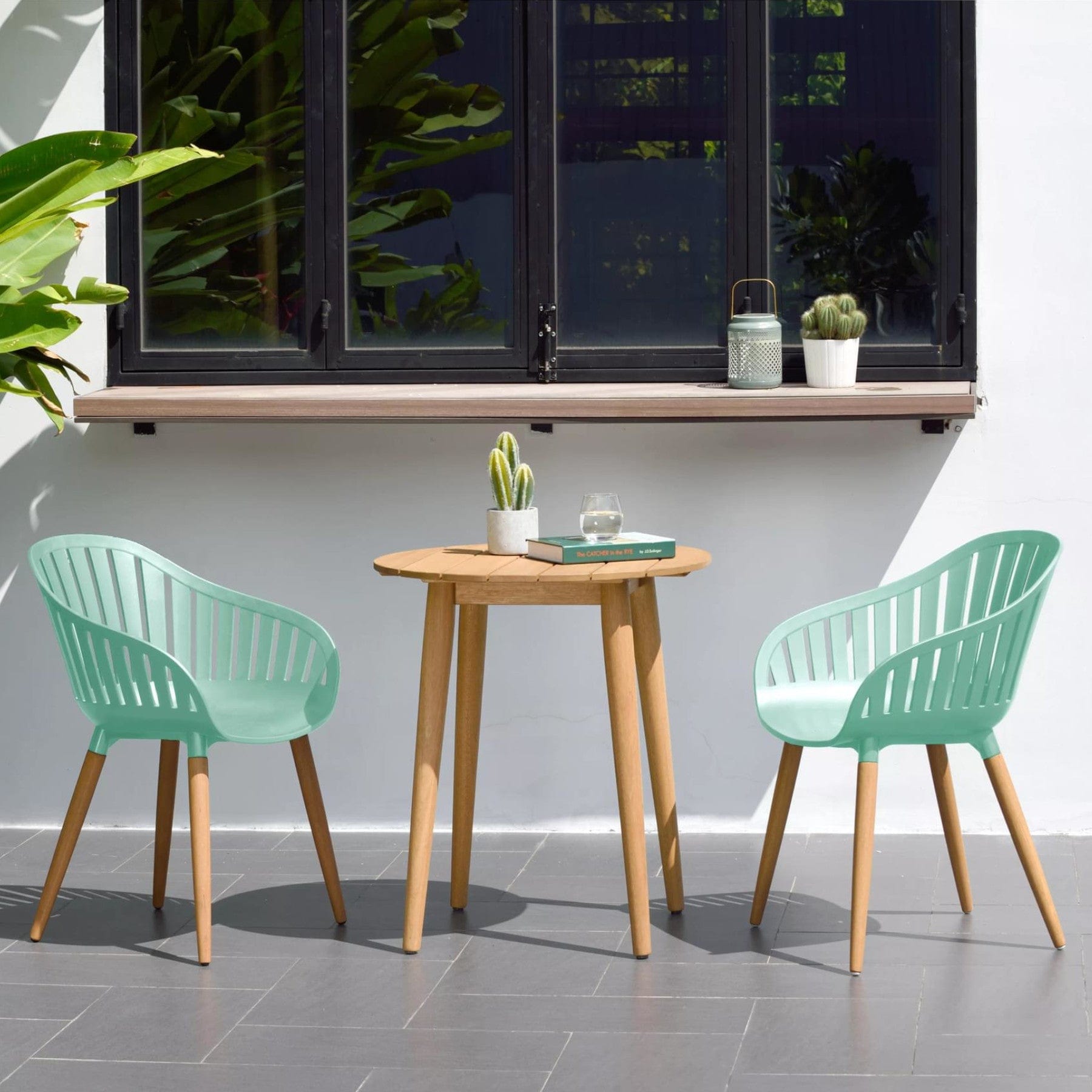 Outdoor patio setup with a round wooden table, two mint green modern chairs, potted cacti, open window, and lush greenery in a tranquil home garden.