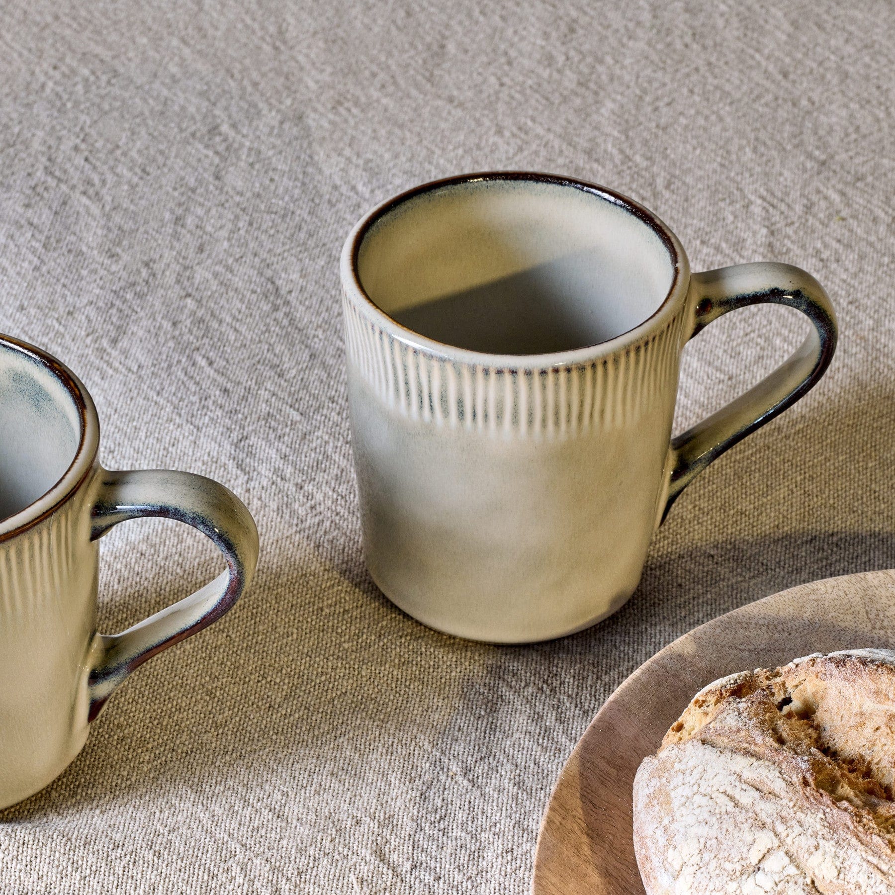 Two ceramic coffee mugs on textured linen tablecloth with rustic homemade bread on wooden plate, cozy breakfast setup, kitchenware, artisanal pottery and bakery concept.