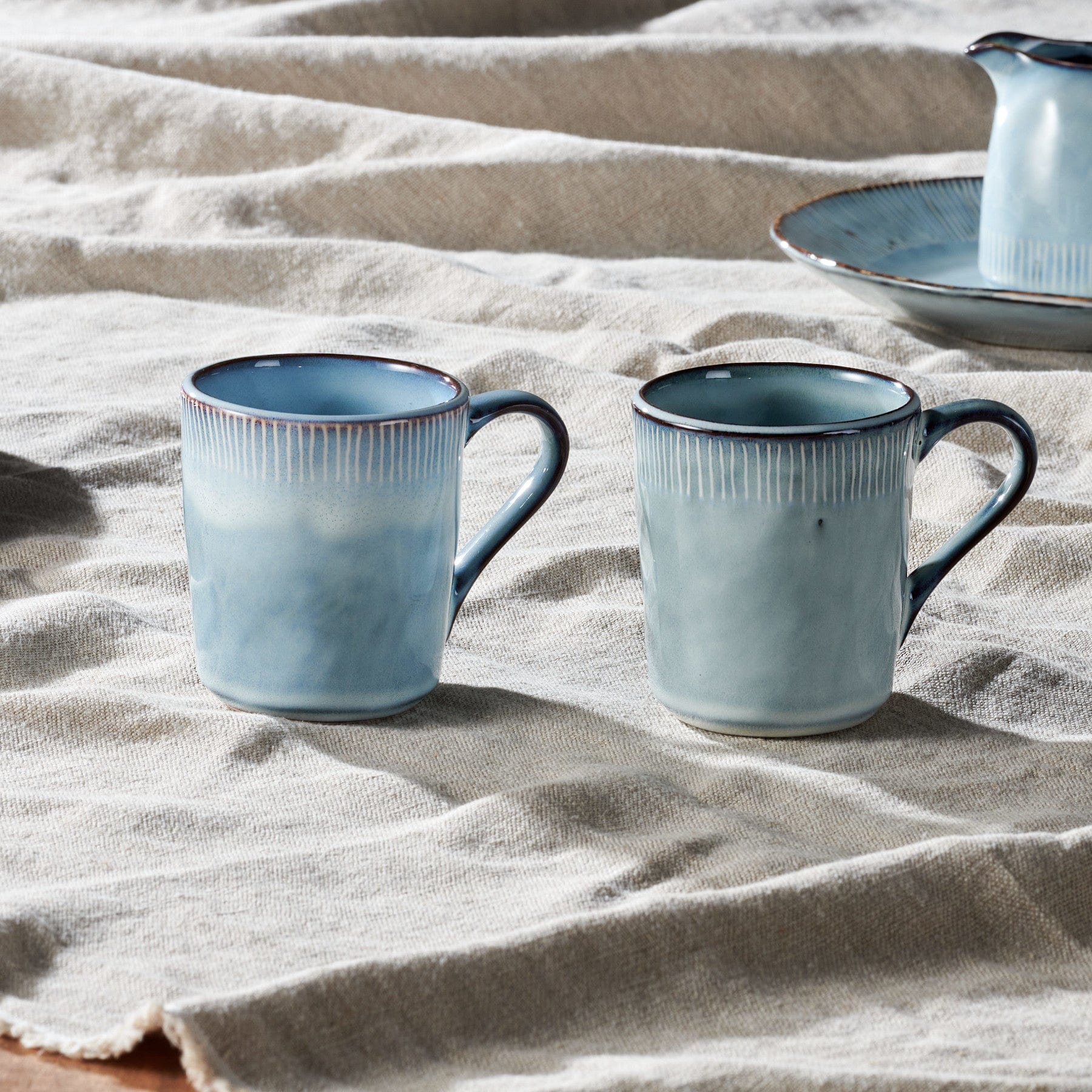 Two blue ceramic coffee mugs on beige linen tablecloth with sunlight casting soft shadows, artisan pottery, cozy home breakfast table setting, handmade stoneware cups with unique glaze texture.