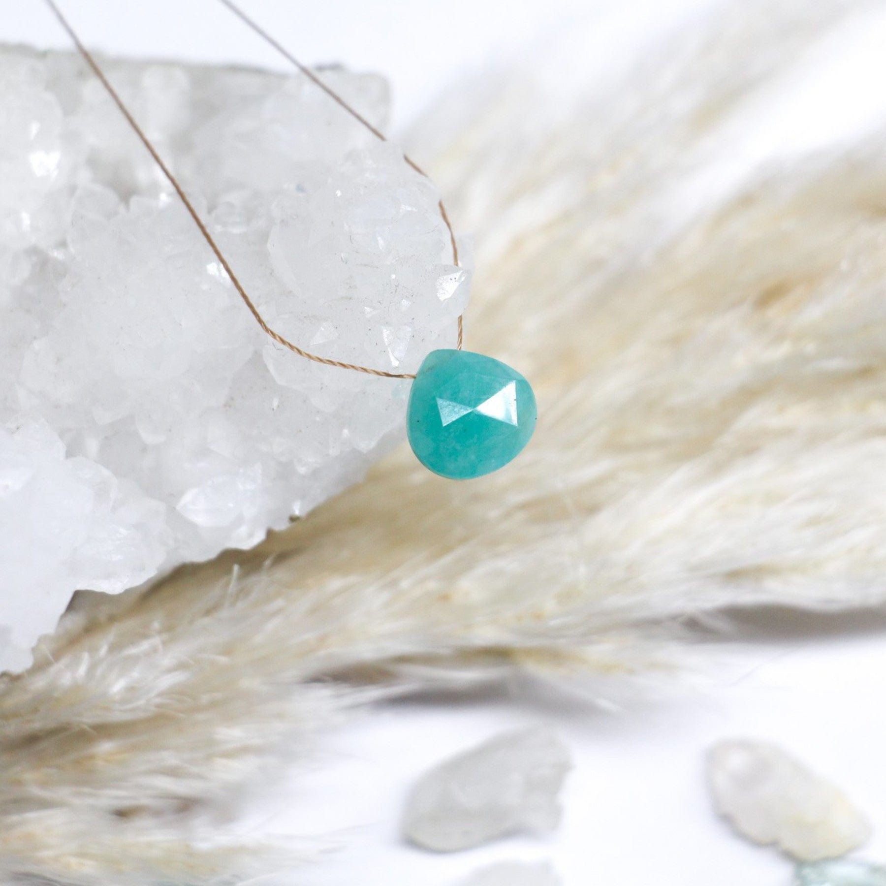 Turquoise gemstone pendant on thin chain over crystal geode, with soft white background and feather texture accents.