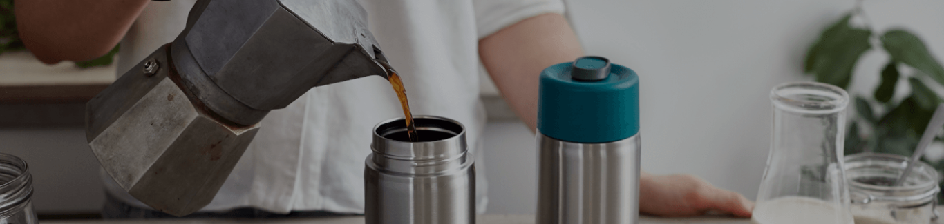 Person pouring coffee from moka pot into stainless steel insulated travel mug on kitchen counter alongside milk bottle and sugar container.