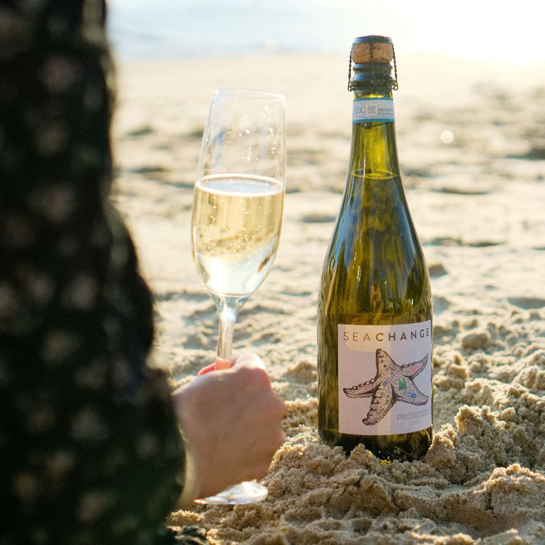 Close-up of champagne glass and bottle of Seachange Prosecco on sandy beach with person's hand, sunset lighting, celebratory beachside drink.