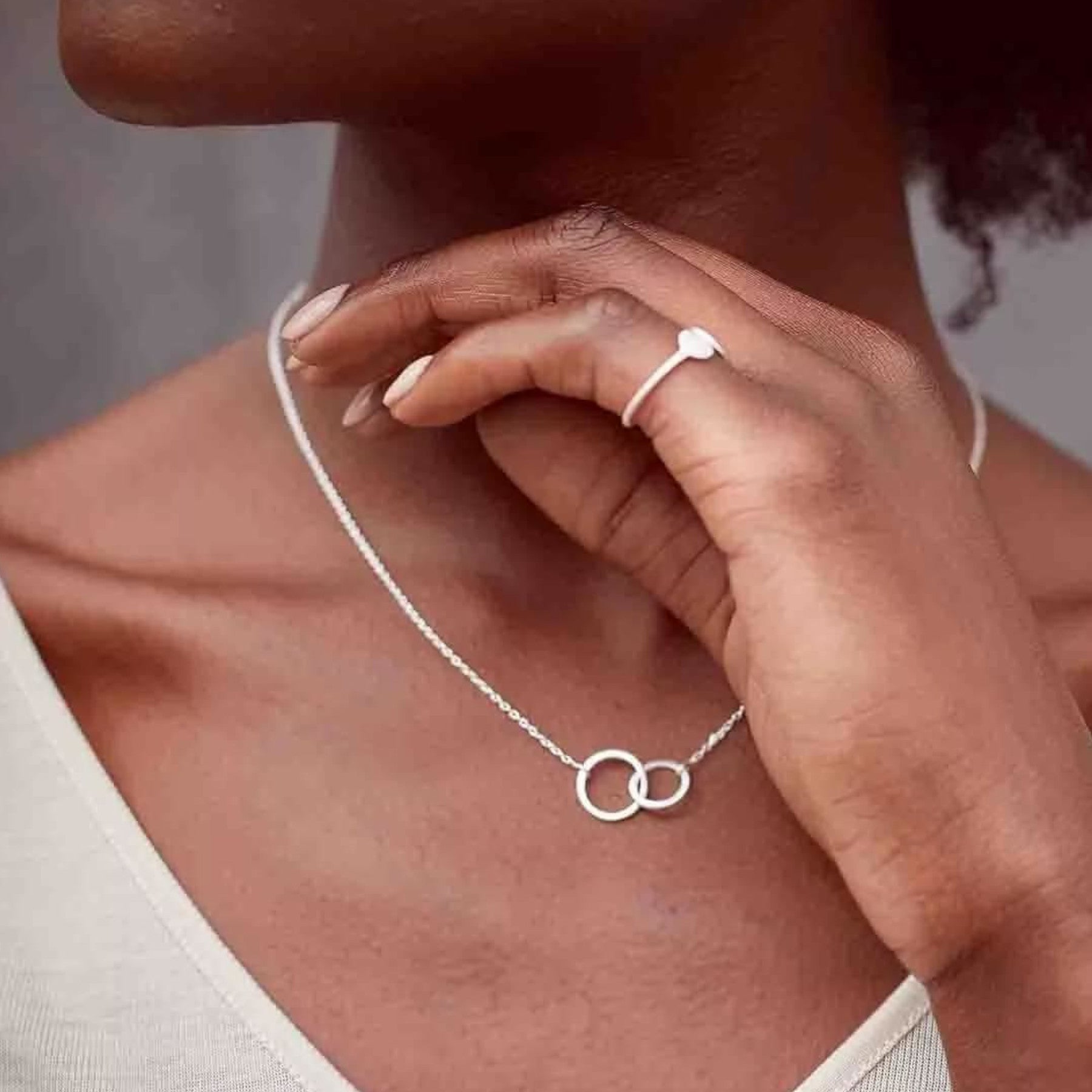 Woman displaying silver necklace with interlocking circles pendant, elegant minimalist jewelry, subtle tan dress, manicured fingernails, fashion accessories, close-up of neck and chest area.