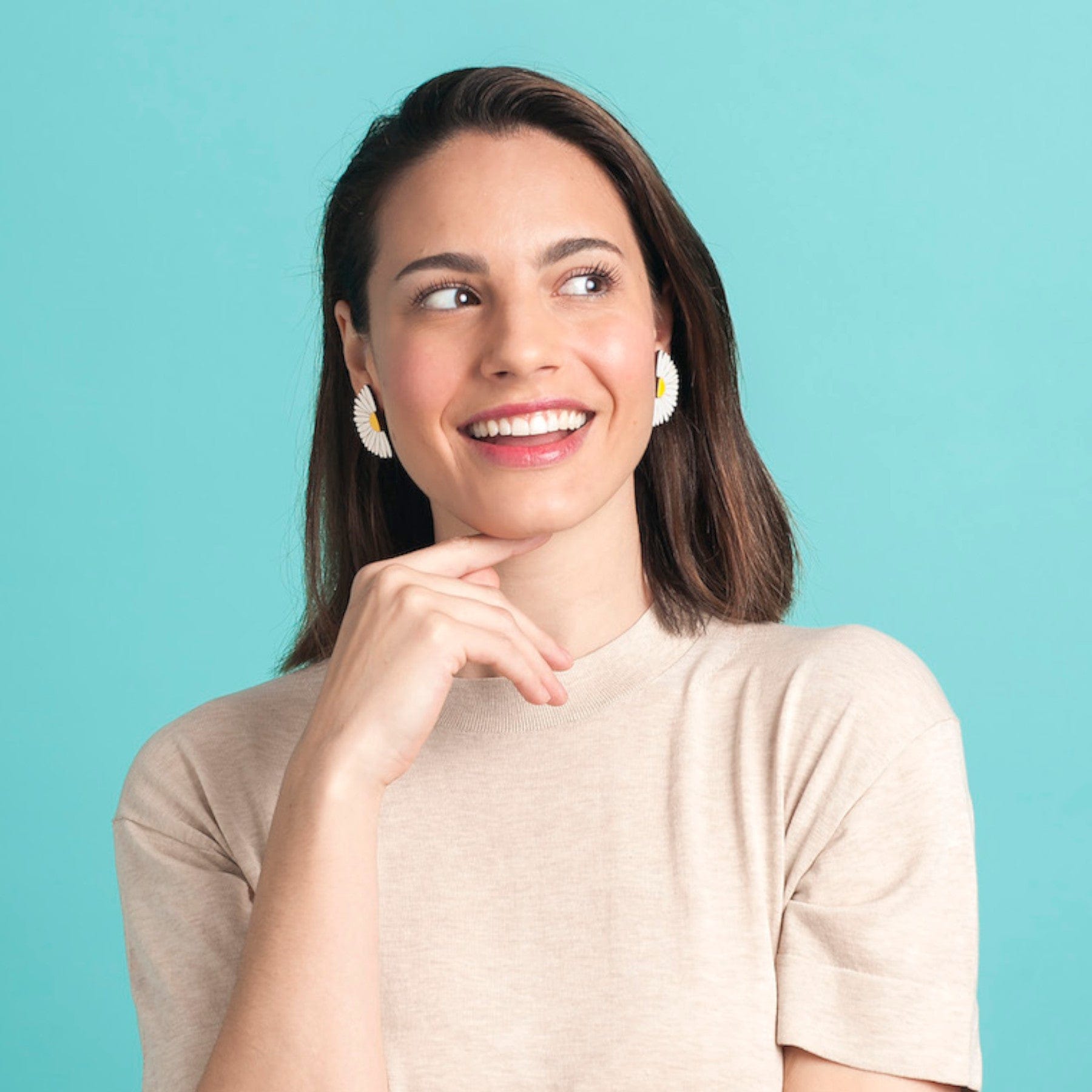 Smiling young woman with daisy earrings looking away thoughtfully against a teal background, wearing a beige top.