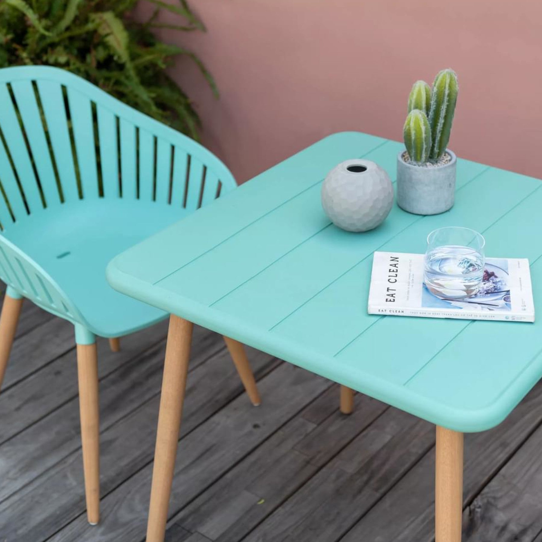 Modern outdoor patio with mint green chair and coordinating table, geometric vase, potted cactus, glass of water, and 'Eat Clean' magazine, against a pink wall on a wooden deck.